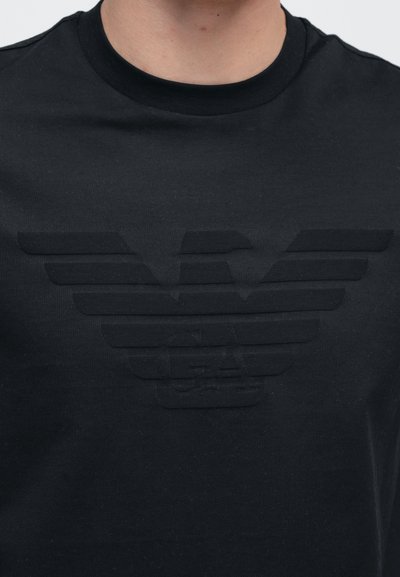 Μαύρο βαμβακερό T-shirt με ανάγλυφο λογότυπο που απεικονίζει στυλιζαρισμένα φτερά. Λαιμόκοψη τύπου crew και κοντά μανίκια. Απλός, μινιμαλιστικός σχεδιασμός.