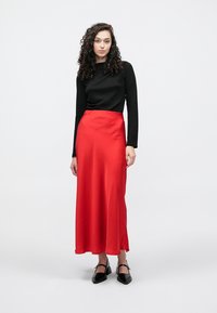 Zwarte top met lange mouwen gecombineerd met een vloeiende rode satijnen rok met een zijsplit. Model draagt zwarte puntige platte schoenen. Eenvoudig, elegant ontwerp.