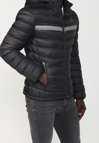 Black puffer jacket with a hood, featuring horizontal stitching, a gray accent stripe, and zippered side pockets. Material appears shiny and smooth.