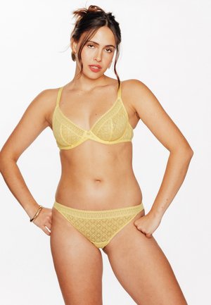 Femme aux cheveux foncés attachés en chignon, portant un soutien-gorge en dentelle jaune et des sous-vêtements assortis, debout les mains sur les hanches devant un fond blanc.