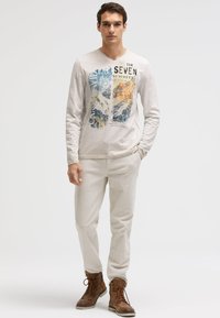 Hellgraues Langarmshirt mit Berggrafik und Text, kombiniert mit hellbeigen Hosen und braunen Schnürstiefeln. Lässiges Outfit dargestellt.