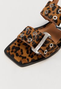 Sandales plates imprimées léopard avec deux larges brides ornées de quincaillerie en argent, un bout carré, et un matériau texturé ressemblant à de la fourrure.