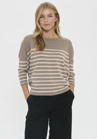 Donna con lunghi capelli biondi che indossa un maglione a righe taupe e bianche e pantaloni neri, in piedi con le mani in tasca contro uno sfondo semplice.