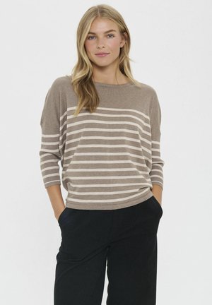 Žena s dlhými blond vlasmi, oblečená v svetlohnedom a bielom pruhovanom svetri a čiernych nohaviciach, stojí so založenými rukami v kapsách pred jednoliatym pozadím.