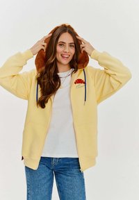 Žltá mikina na zips s kapucňou vystlanou hnedou, vyšívaným logom croissantu. Má modré šnúrky a dve predné vrecká.