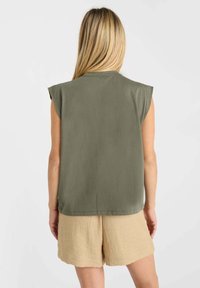 Femme aux longs cheveux blonds portant un top sans manches vert olive et un short beige texturé, se tenant de dos devant un fond uni.