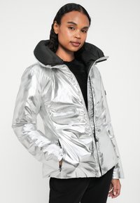 Casaco acolchoado prateado com um acabamento brilhante, featuring capuz e fecho de correr pretos, e detalhes franzidos. O tecido texturizado acrescenta interesse visual.