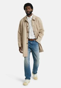 Helle beige Trenchcoat mit Knopfleiste, über einem weißen Hemd und blauen Jeans getragen. Die Person trägt cremefarbene Sneaker und steht in einer neutralen Pose.