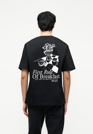 Άτομο που φοράει μαύρο μπλουζάκι με λευκό γραφικό που απεικονίζει κανάτα καφέ, φλιτζάνι και τοστ σε τραπέζι με καρό μοτίβο και κείμενο σχετικά με το πρωινό.