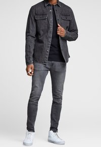 Mann in schwarzer Jeansjacke, schwarzem Hemd, grauen Jeans, weißen High-Top-Sneakern und Ringen an den Fingern, vor einem einfarbigen Hintergrund stehend.