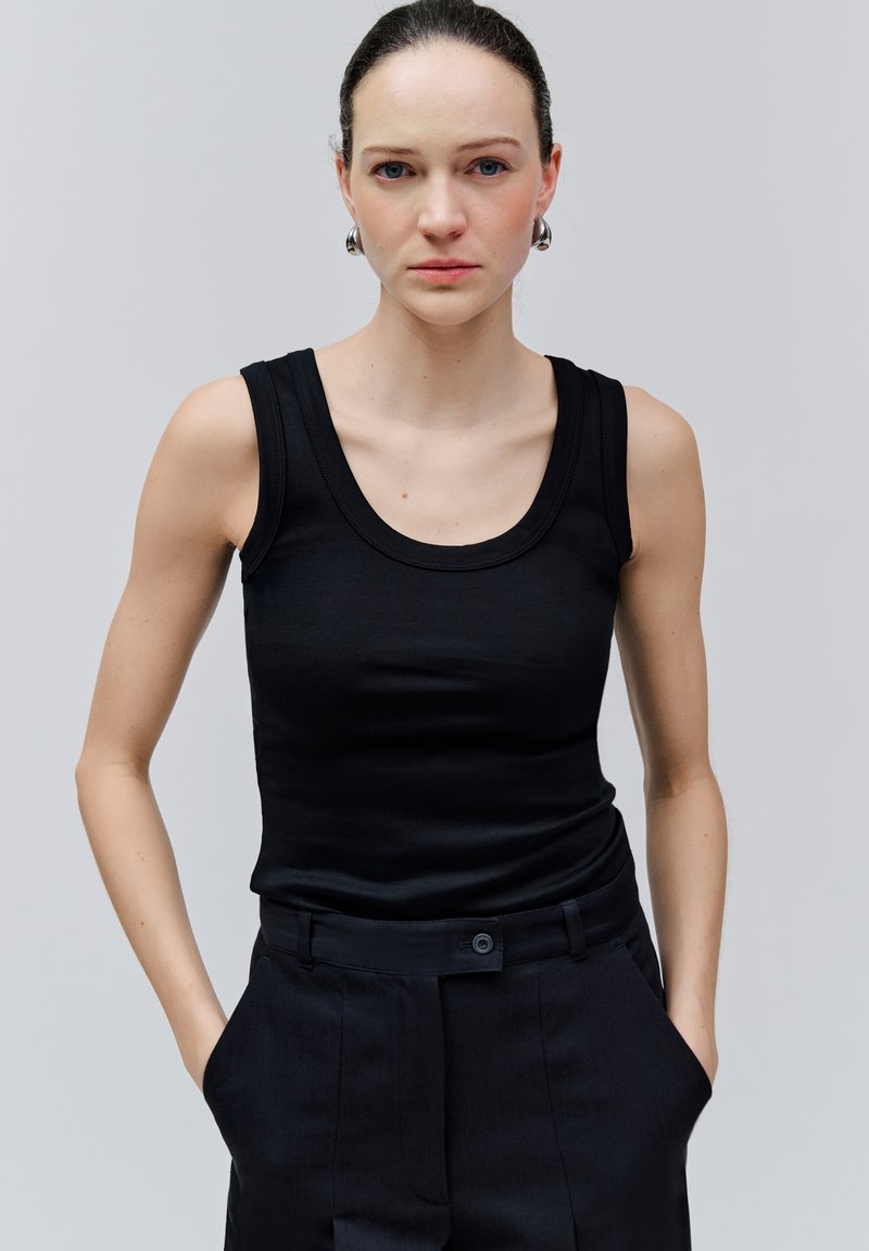 Jonge vrouw met een lichte huid en blauwe ogen, gekleed in een zwart mouwloos topje en zwarte broek, staande met handen in haar zakken tegen een effen achtergrond.