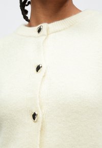 Cardigan de couleur crème en matière douce et texturée, avec un col rond et quatre boutons en métal brillant.