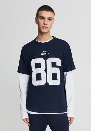 Maglietta di cotone blu marino con maniche corte, sotto una maglia a maniche lunghe bianca a strati, grande stampa bianca testurizzata con il numero "86" e testo "NYC ATHLETIC" sopra.