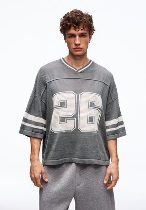 Junger Mann mit lockigem Haar, der ein übergroßes graues Sporttrikot mit der Nummer 26 und passende graue Jogginghosen trägt, vor weißem Hintergrund stehend.