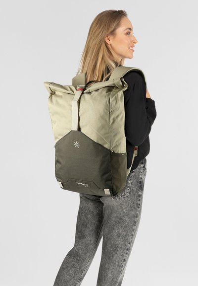 Donna bionda in camisa nera e jeans grigi che indossa uno zaino a rullino beige e verde scuro, sorridente e che guarda di lato su uno sfondo bianco.