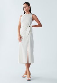 Mouwloze beige jurk met een gestrikte taille, een kniehoogte split aan de voorkant en een gestructureerde stof. Gecombineerd met witte open-toe hakken.