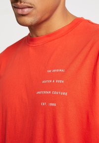 Close-up van een persoon die een rood t-shirt draagt met witte tekst waarop staat "The Original Scotch & Soda Amsterdam Couture Est. 1985" op de borst.