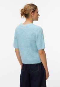 Ženska s svetlimi lasmi zavezano v figo, oblečena v svetlo modro puhasto kratko majico z rokavi in temne široke kavbojke, obrnjena stran od kamere.