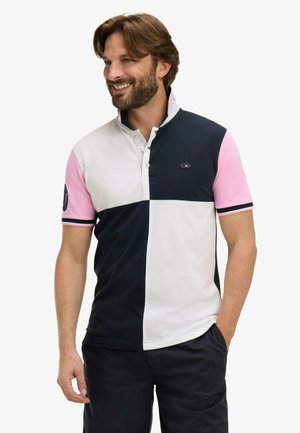 Homme souriant, portant un polo bicolore avec des sections bleu marine, blanc et rose, debout les mains dans la poche, sur un fond uni.