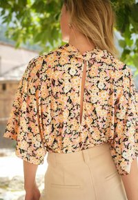 Blomstermønstret bluse med korte ærmer, der har en nøglehul-åbning bagpå. Farverne inkluderer sort, pink, creme og orange. Letvægtsstof.