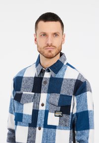 Camicia di flanella con una fantasia a quadri blu e bianca, dotata di chiusure a bottoni, due tasche sul petto e un logo a patch. Realizzata in tessuto morbido.