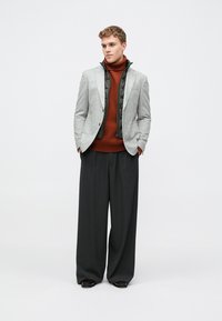 Grauer Blazer über einer schwarzen gesteppte Weste, kombiniert mit einem rostfarbenen Rollkragenpullover und lässigen schwarzen Hosen. Glattes Material mit taillierter Passform.