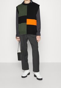 Chaleco de punto con bloques de color verde, negro y naranja, llevado sobre una camisa oscura. Combinado con pantalones grises y zapatos de plataforma blancos. Bolso negro.