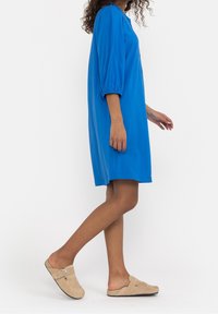 Abito blu al ginocchio con maniche a sbuffo, texture liscia e vestibilità comoda. Abbinato a scarpe beige slip-on con un accento circolare.