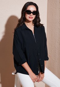 Camicia nera con bottoni e maniche arrotolate, realizzata in tessuto leggero. Abbinata a pantaloni bianchi. Modello con occhiali da sole neri.
