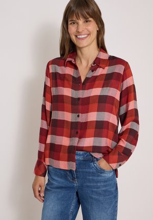 Mujer con una camisa de cuadros roja y rosa abotonada y vaqueros azules, sonriendo con una mano en el bolsillo.