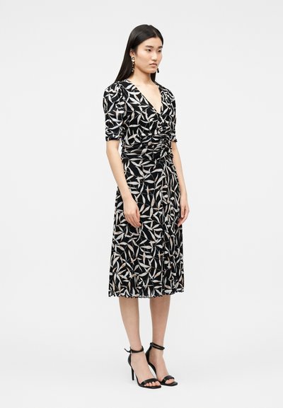 Γυναίκα που φοράει μαύρο μεσοφόρεμα με λευκά και μπεζ σχέδια φύλλων, με πιέτες στο μπροστινό μέρος, φουσκωτά μανίκια και μαύρες ψηλοτάκουνες γόβες με λουράκι στον αστράγαλο, στέκεται πλαγίως.