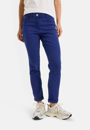 Personne portant un pantalon bleu retroussé, des baskets blanches et une chemise blanche, debout devant un fond uni.