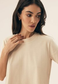 Jasno beżowa koszula w paski z okrągłym dekoltem i krótkimi rękawami, wykonana z miękkiego materiału, z delikatną fakturą i luźnym krojem.