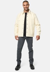 Beige Cord-Jacke mit Druckknöpfen, zwei Fronttaschen, kombiniert mit einem schwarzen T-Shirt und grauen Jeans. Schwarze Stiefel runden das Outfit ab.