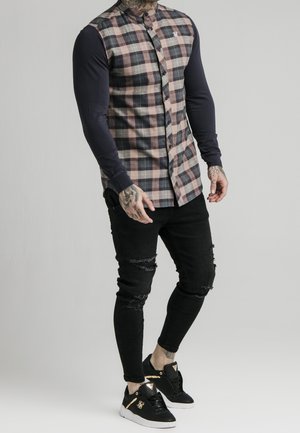 Mężczyzna w kraciastej koszuli z krótkim rękawem nałożonej na czarną koszulę z długim rękawem, podartych czarnych dżinsach i czarnych trampkach z białymi podeszwami.
