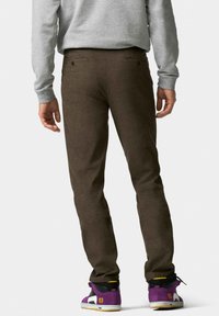 Bruine broek van gestructureerde stof, met achterzakken en een slim fit. Draagt een paar paarse en zwarte sneakers.