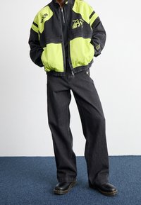 Svart och neongrön jacka med dragkedja och logodetaljer, kombinerad med svarta vida byxor och svarta skor på en blå matta.