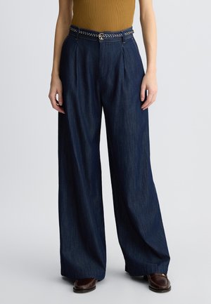 Pantaloni in denim blu scuro a gamba larga con una cintura intrecciata, caratterizzati da una vita alta e pieghe frontali, abbinati a scarpe brown slip-on.