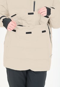 Beige isolerad jacka med dragkedja och kardborre-lommer; har elastiska ärmslut och hög krage. Synlig sömnad och jämn tygtextur.