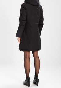 Abrigo de invierno negro, hasta las rodillas, con capucha, que presenta mangas largas y una tela suave y texturizada. Acompañado de botas hasta el tobillo.