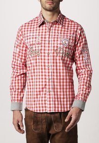 Camisa de cuadros rojos y blancos con mangas largas, cierre delantero de botones y puños grises. Presenta acentos bordados y parches decorativos.