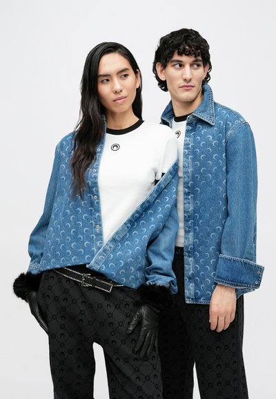 Vaaleansininen kuvioitu denimtakki, yhdistetty valkoiseen paitaan ja mustiin kuvioituihin housuihin. Mustat hansikkaat ja niitattu vyö täydentävät asua.