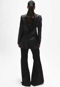 Kobieta z długimi ciemnymi włosami związanymi w kucyk, ubrana w czarną skórzaną marynarkę i szerokie czarne spodnie, stojąca tyłem na białym tle.