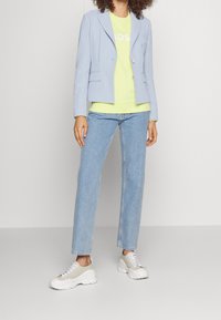 Blazer azzurro chiaro sopra una felpa verde neon, abbinato a jeans azzurri chiari e sneaker bianche chunky, con lacci e dettagli grigi.