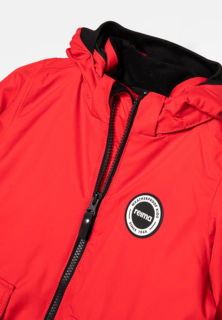 Chaqueta impermeable roja para niños con cremallera negra, capucha ajustable y parche circular blanco con el logo que dice "reima impermeable para niños desde 1944."