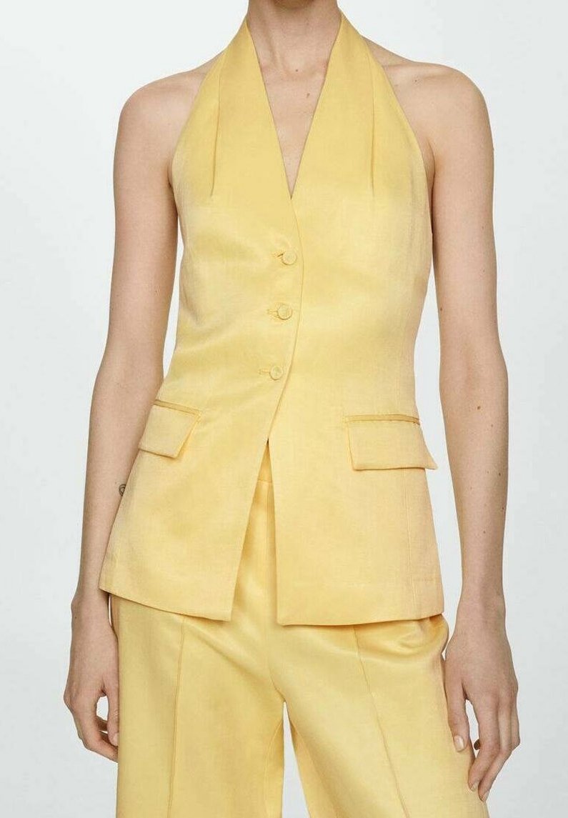 Femme porte un gilet jaune sans manches, taillé sur mesure, avec trois boutons et un pantalon large taille haute assorti, sur fond uni.