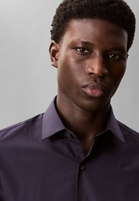 Dark purple button-up shirt with a collar, smooth texture, and subtle sheen. Model's face features strong jawline and natural makeup.