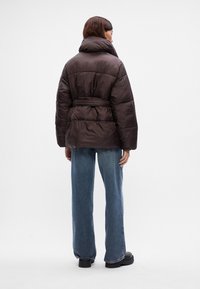 Smeđi puffer jakna s visokim ovratnikom, pojasom u struku i oversized krojem. Ima prošivenu teksturu i široke rukave. Nosena s plavim trapericama.