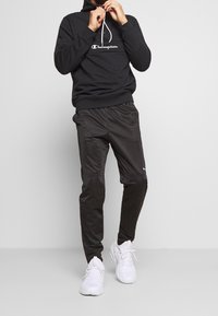 Felpa con cappuccio nera con logo bianco, abbinata a pantaloni sportivi neri a forma affusolata e sneaker bianche. Texture liscia per tutto l'insieme.