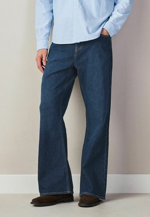 Mann trägt dunkelblaue, weit geschnittene Jeans, braune Schuhe und ein hellblau gestreiftes Langarmhemd, eine Hand in der Tasche.